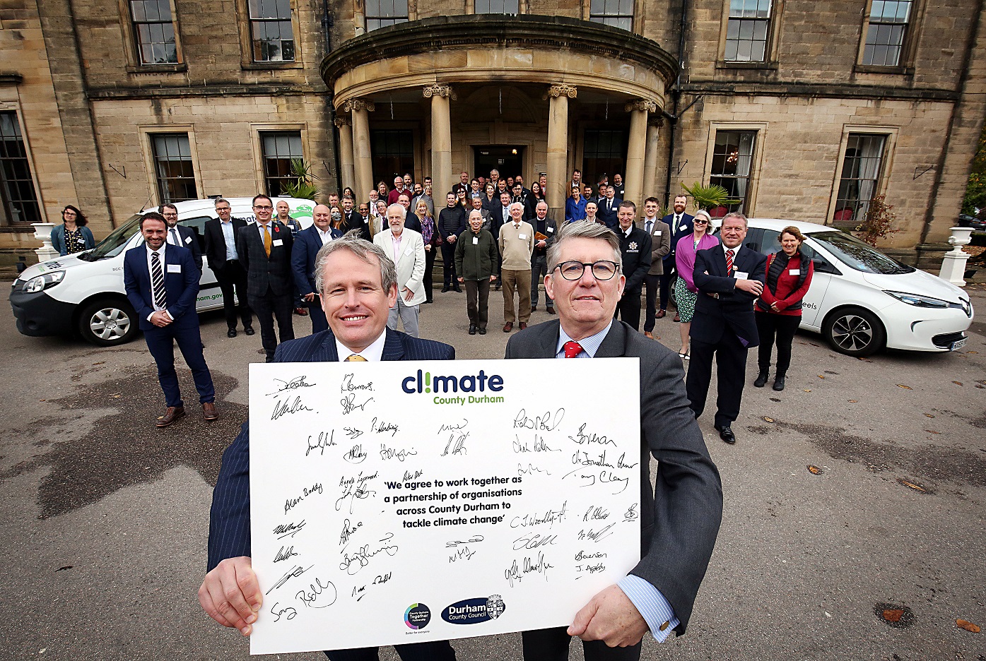 Two people at forefront of picture holding Climate County Durham pledge including lots of signatures of those many people stood in the background. Backdrop is Beamish Hall and two electric vehicles.