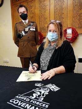 woman sat at table signing document with a man in uniform stood behind